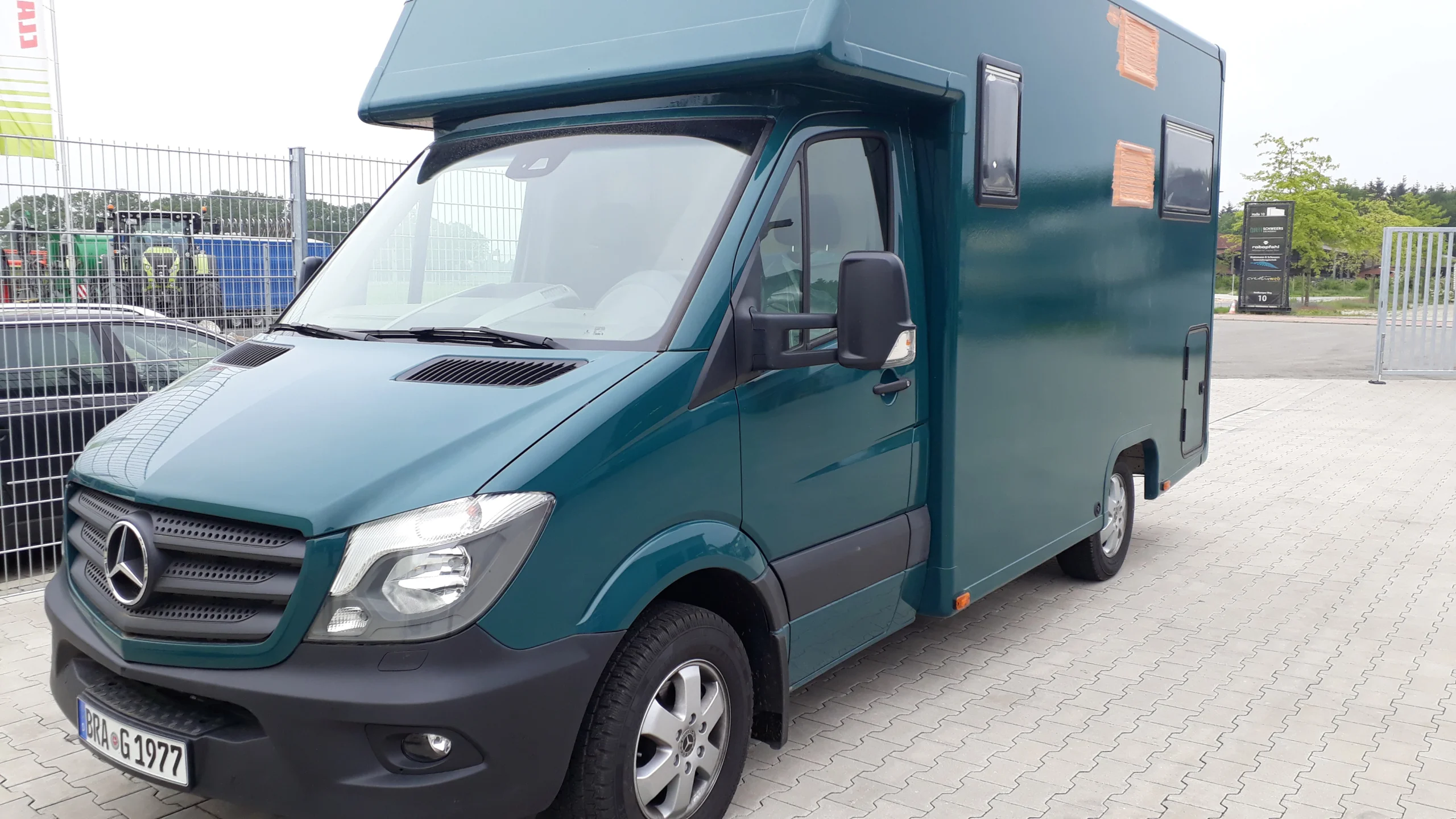 Grüner Mercedes-Sprinter Campingbus von vorne rechts auf gepflastertem Parkplatz, seitliche Fenster und Tür sind sichtbar, Hinterhof mit Zaun und Fahrzeugen im Hintergrund