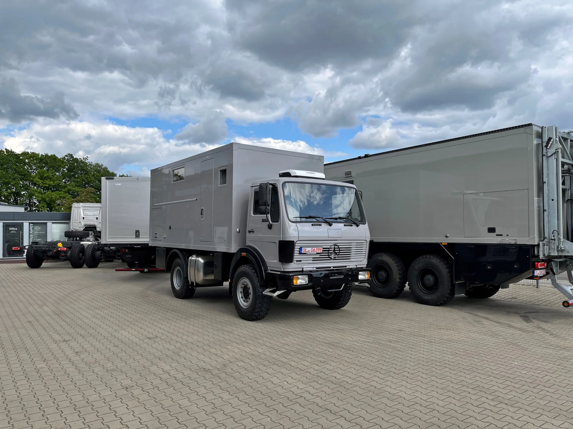 Mehrere graue Lastwagen mit großen Aufbauten, die auf einem gepflasterten Parkplatz unter stark bewölktem Himmel geparkt sind, im Hintergrund ein Gebäude mit Bäumen.