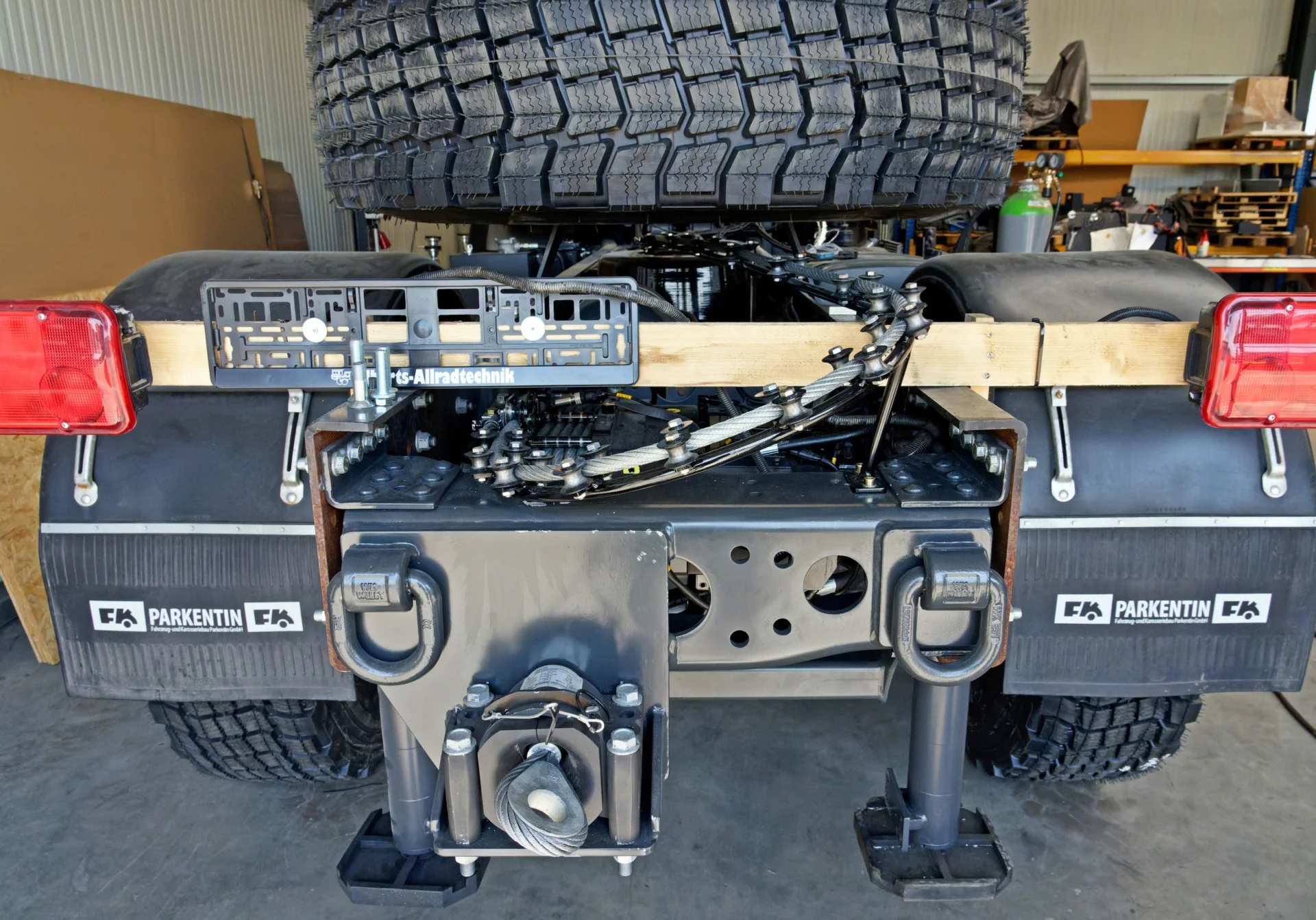 Heckansicht eines robusten Fahrgestells eines Geländefahrzeugs in Werkstatt, mit großem Reserverad, zwei roten Rücklichtern, Anhängerkupplung, Metallrahmen und zwei großen Reifen im Hintergrund.