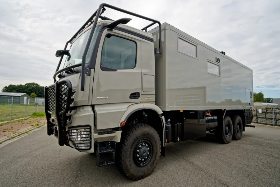 Grau lackierter Expeditions-LKW mit großem, ausgebautem Heckaufbau auf Asphaltparkplatz vor leicht bewölktem Himmel und eingezäuntem Gelände im Hintergrund.