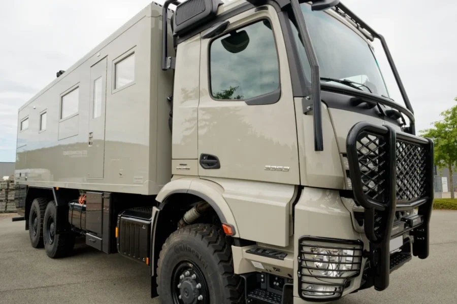 Beige schwerer Allrad-LKW mit einem großen Aufbau und schützenden schwarzen Metallgittern vor Frontscheibe und Scheinwerfern, geparkt auf Asphalt mit Bäumen im Hintergrund