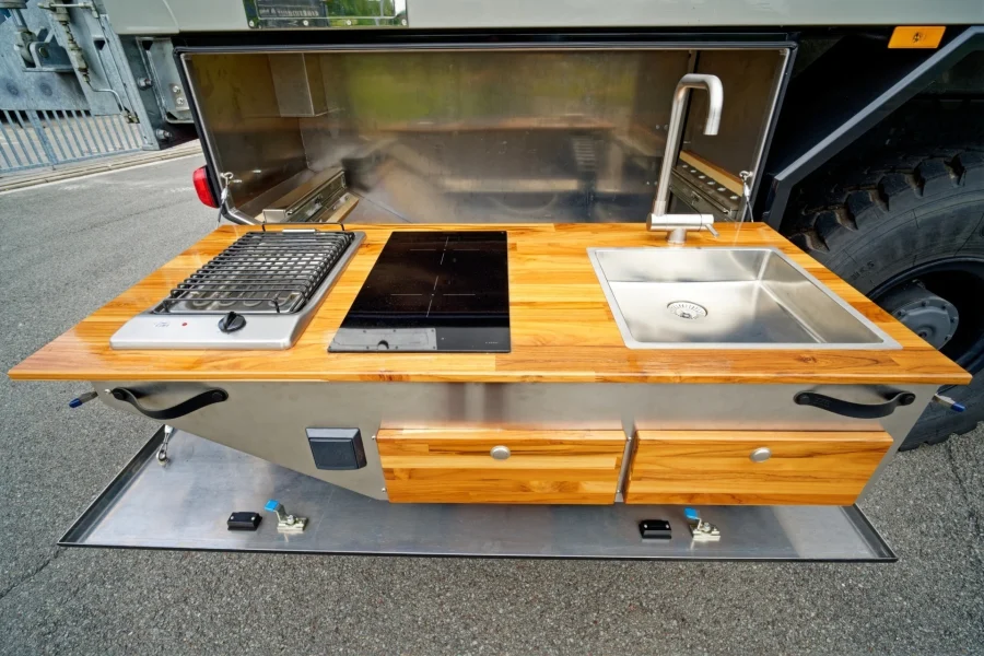Ausziehbare Campingküche mit Holzarbeitsplatte, Gasofen, Induktionskochfeld und Edelstahlspüle mit Wasserhahn, integriert in ein Fahrzeug mit großem Reifen im Hintergrund.