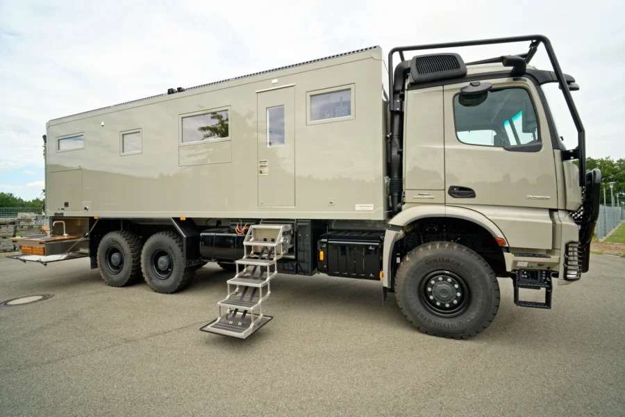Beige Expeditions-Lkw mit ausgefahrener Trittleiter und mehreren Fenstern, geparkt auf asphaltierter Fläche, im Hintergrund ein Zaun und Bäume.