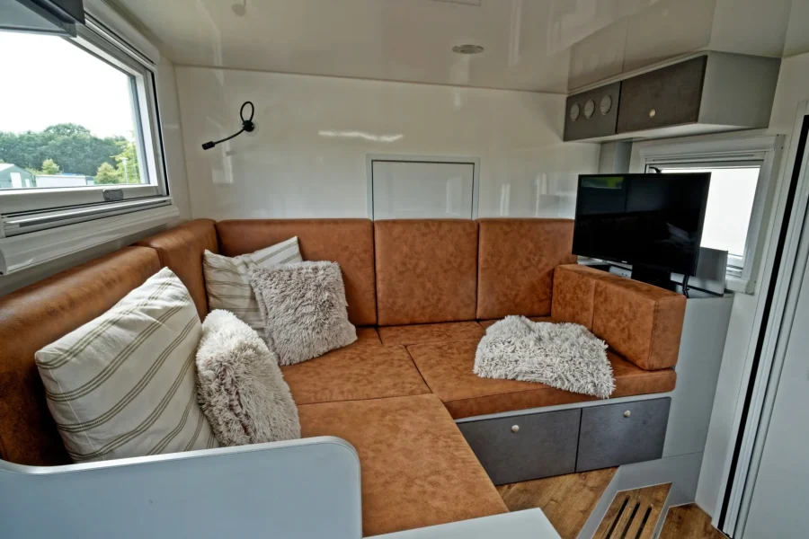 Innenraum eines Wohnmobils mit braunem Ecksofa, mehreren Kissen in Beige- und Grautönen, einem kleinen Fenster an der linken Seite und einem Fernseher an der rechten Seite, weißen Wänden und Holzfußboden.