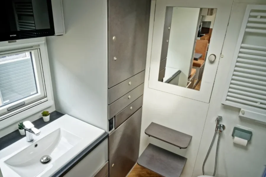 Kompaktes Badezimmer mit Holzfußboden, Waschbecken mit Unterschrank, hohen Schränken, Toilette, Spiegel an der Wand, Handbrause und Blick auf den Innenraum durch eine Fensterklappe in der Decke.