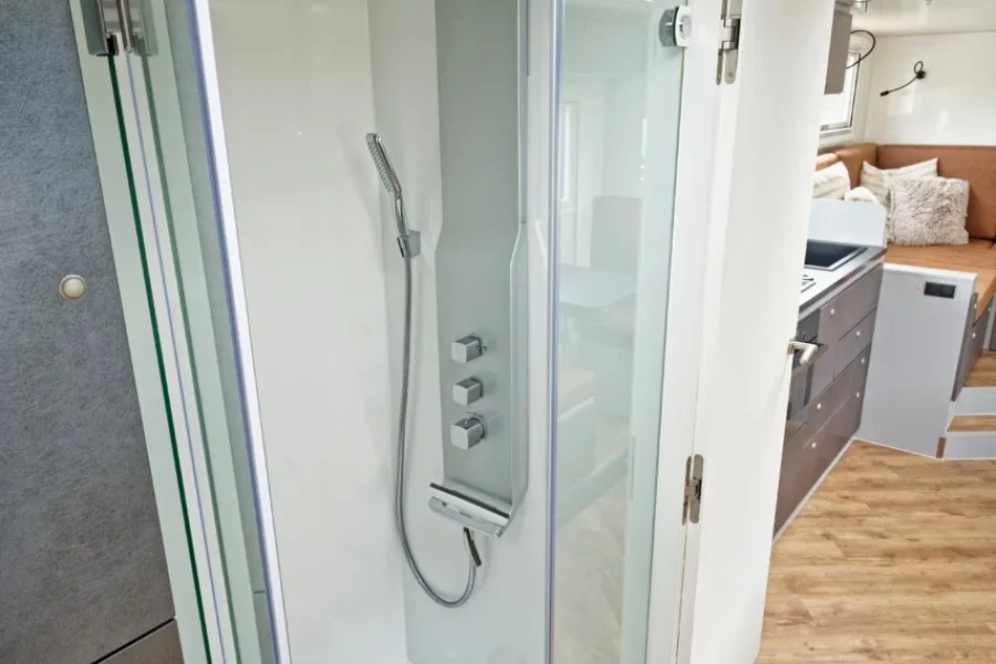 Kleine moderne Dusche mit Glasabtrennung in einem kompakten Wohnmobil oder kleinem Apartment, Blick auf angrenzenden Raum mit Küchenzeile und Sitzbereich.