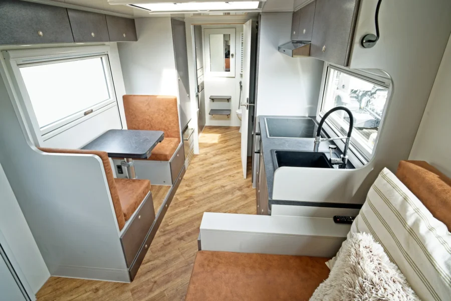 Innenansicht eines modernen Wohnmobils mit braunen Sitzbänken, einem kleinen Esstisch links, einer Küche mit Spüle und Herd rechts sowie Blick auf den hinteren Bereich mit offenem Badzimmer und Spiegel.