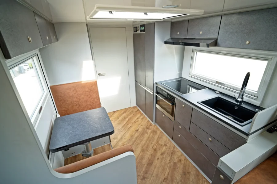 Innenraum einer modernen Wohnmobil-Küche mit grauen Schränken, Kochfeld, Spüle, Backofen, braunen Sitzbänken und einem kleinen Esstisch mit schwarzer Tischplatte, hellem Holzfußboden und einem Fenster über der Arbeitsfläche.