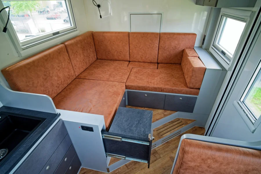 Modern eingerichteter Wohnbereich mit braunem Ecksofa und ausziehbarem, grauem Tisch in einem hellen Raum mit Fenstern und Küchenzeile.