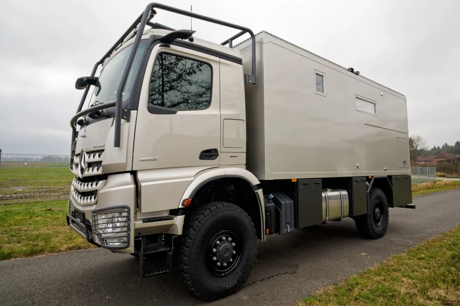 Beige Mercedes-Lkw mit Offroad-Reifen und verstärktem Schutzrahmen, geparkt auf einer Straße neben einem Feld mit Zaun und Gebäuden im Hintergrund bei bewölktem Himmel.