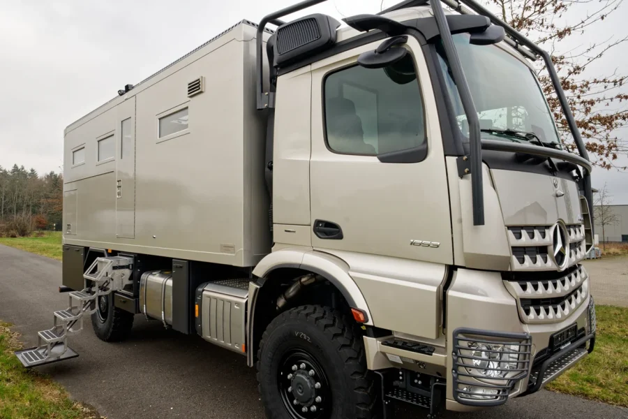 Beiger Expeditions-Lkw mit robuster Kabine und Aufbauten, ausklappbarer Metalltreppe seitlich, steht auf Asphaltstraße bei bewölktem Himmel und Grasflächen im Hintergrund.