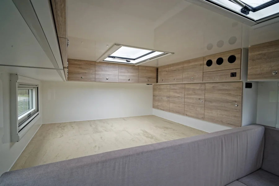 Innenansicht eines hellen, modernen Wohnwagens mit beige Boden, grauem Sofa, Holzfurnierte Schranktüren entlang der Wand und großem Dachfenster in der Decke.
