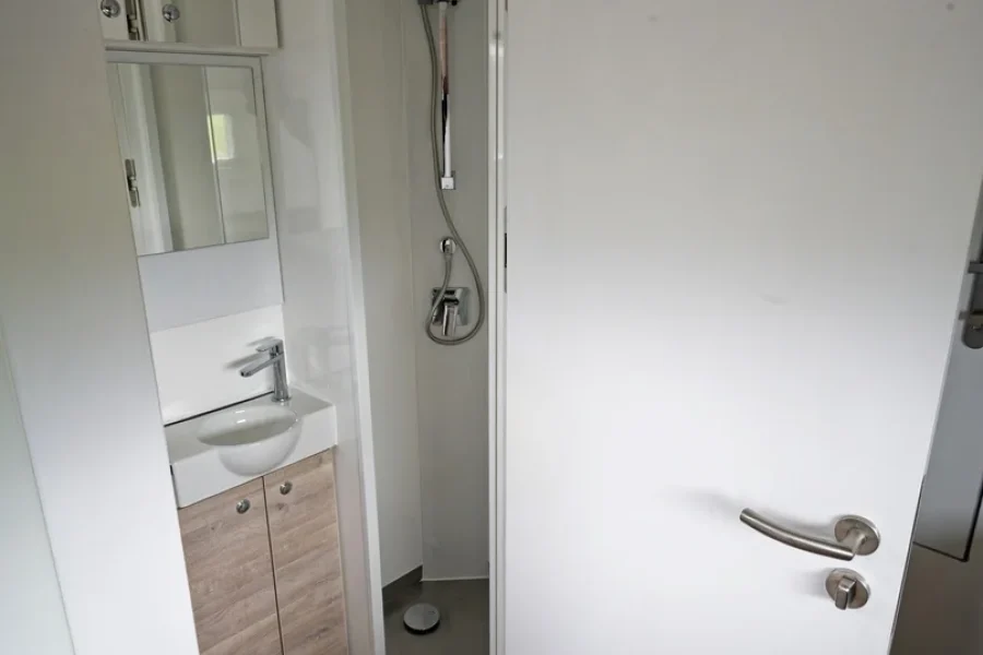 Kleines Bad mit weißen Wänden, einer halb geöffneten weißen Tür, Holzoptik-Boden, einem kleinen Waschbecken mit Unterschrank aus Holzoptik und einem Spiegel mit Schrank darüber sowie einer kleinen offenen Dusche mit verchromtem Duschkopf.