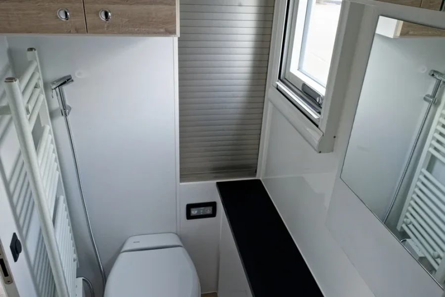 Kompaktes Badezimmer mit weißer Toilette, kleinem Waschbecken mit Chrome-Hahn, Holztäfelung und Teleskop-Duschkopf neben einem Handtuchheizkörper, Fenster mit Jalousie und großem Spiegel an der Wand.