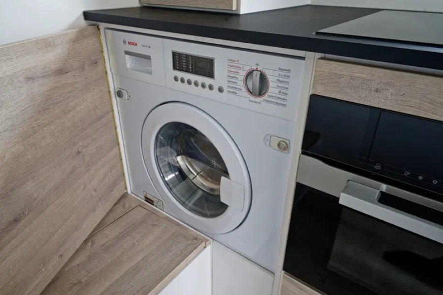 Weiße Bosch Waschmaschine eingebaut in eine moderne Küche mit Holzfronten und schwarzer Arbeitsplatte, daneben ein Backofen von Miele.