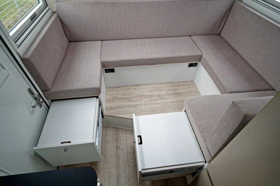 Innenraum eines Wohnmobils mit grauen Polstersitzen in U-Form und geöffnete weiße Schubladen unter den Sitzflächen, Holzfußboden und Fenster mit Sicht nach draußen