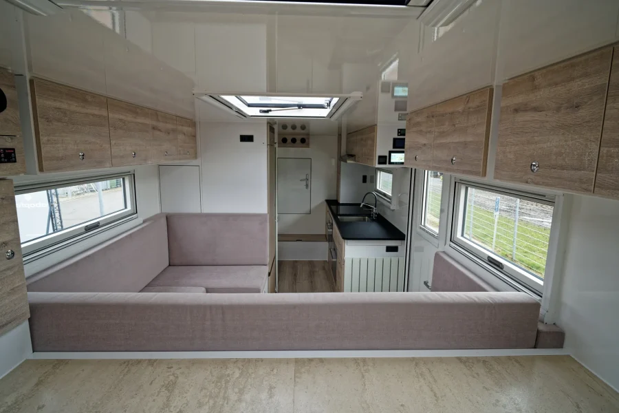 Innenansicht eines modernen Wohnmobils mit Küchenzeile rechts, Sitzbereich links mit hellgrauen Polstern, Holzschränken oberhalb der Fenster und Oberlicht im weißen Deckengehäuse.