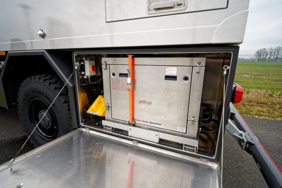 Offenes Fach an einem Fahrzeug zeigt einen Fischer Panda Generator mit Modellbezeichnung 8000x Compact Power, gesichert mit einem orangefarbenen Gurt, neben einem großen Reifen und einer abgesenkten Metalltür.